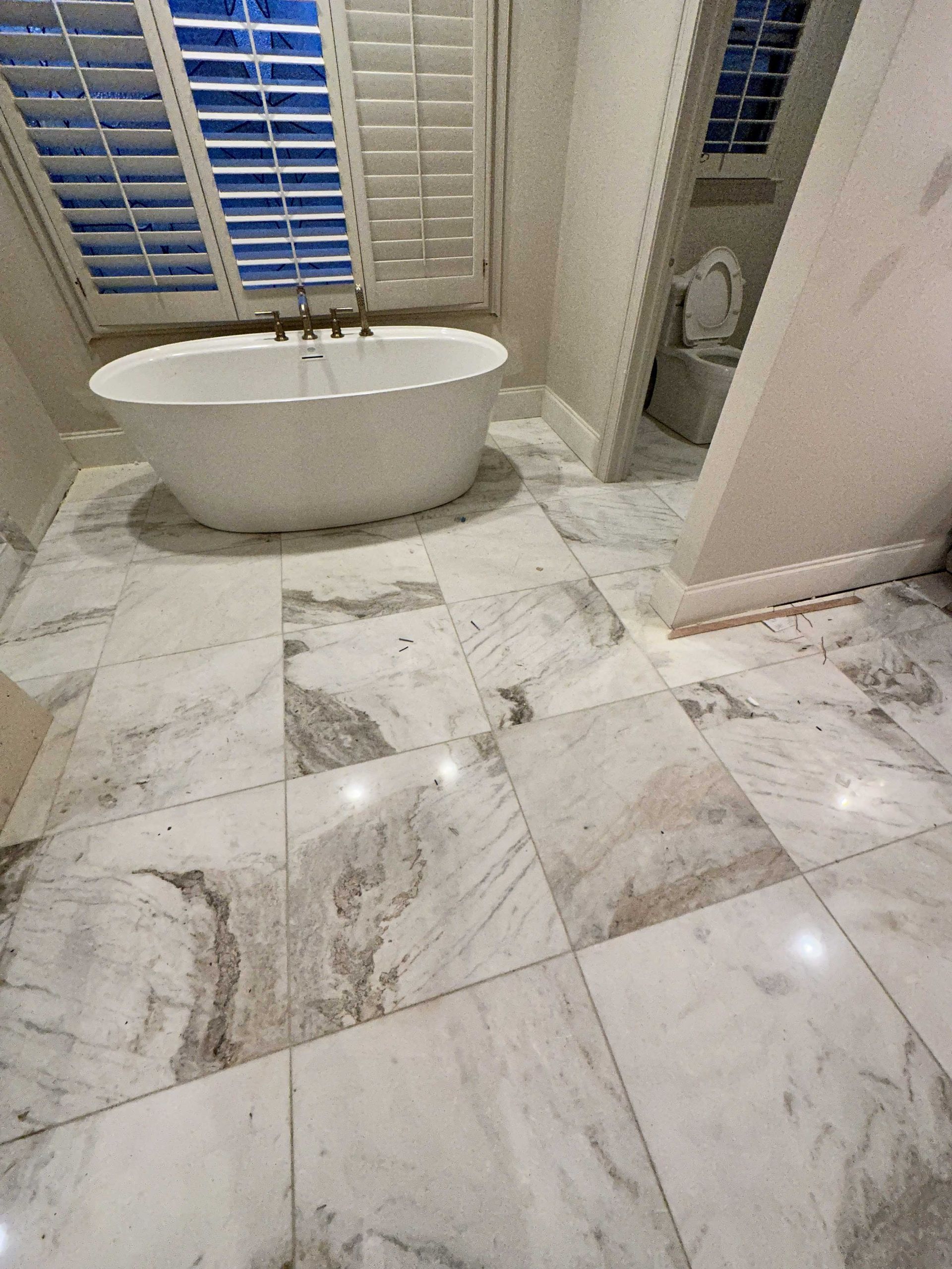Bathroom with a white tub, marble floors, and a toilet. Sunlight streams through window shutters.