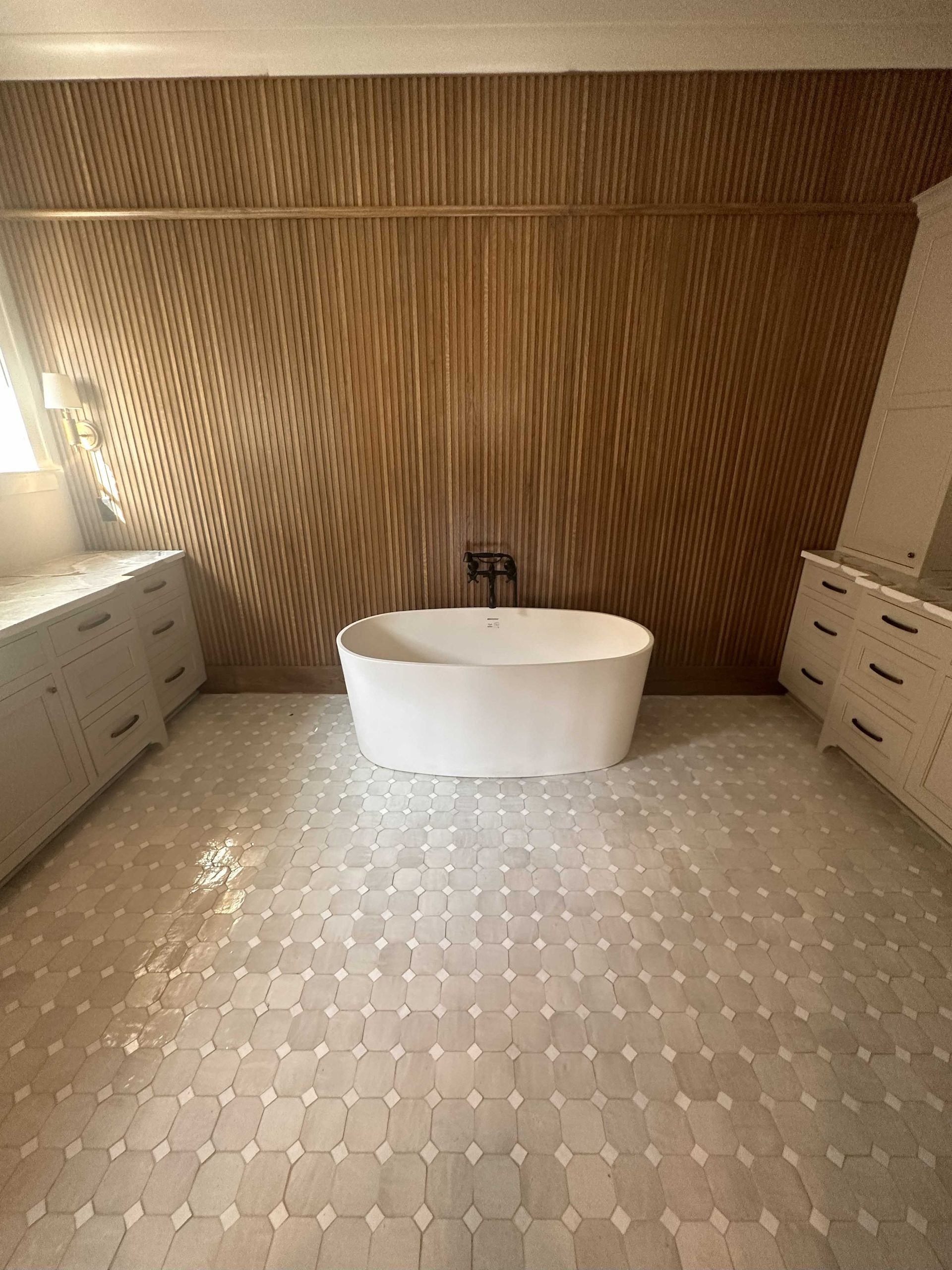 Bathroom with a freestanding tub, wood accent wall, and patterned tile floor. White vanities on either side.