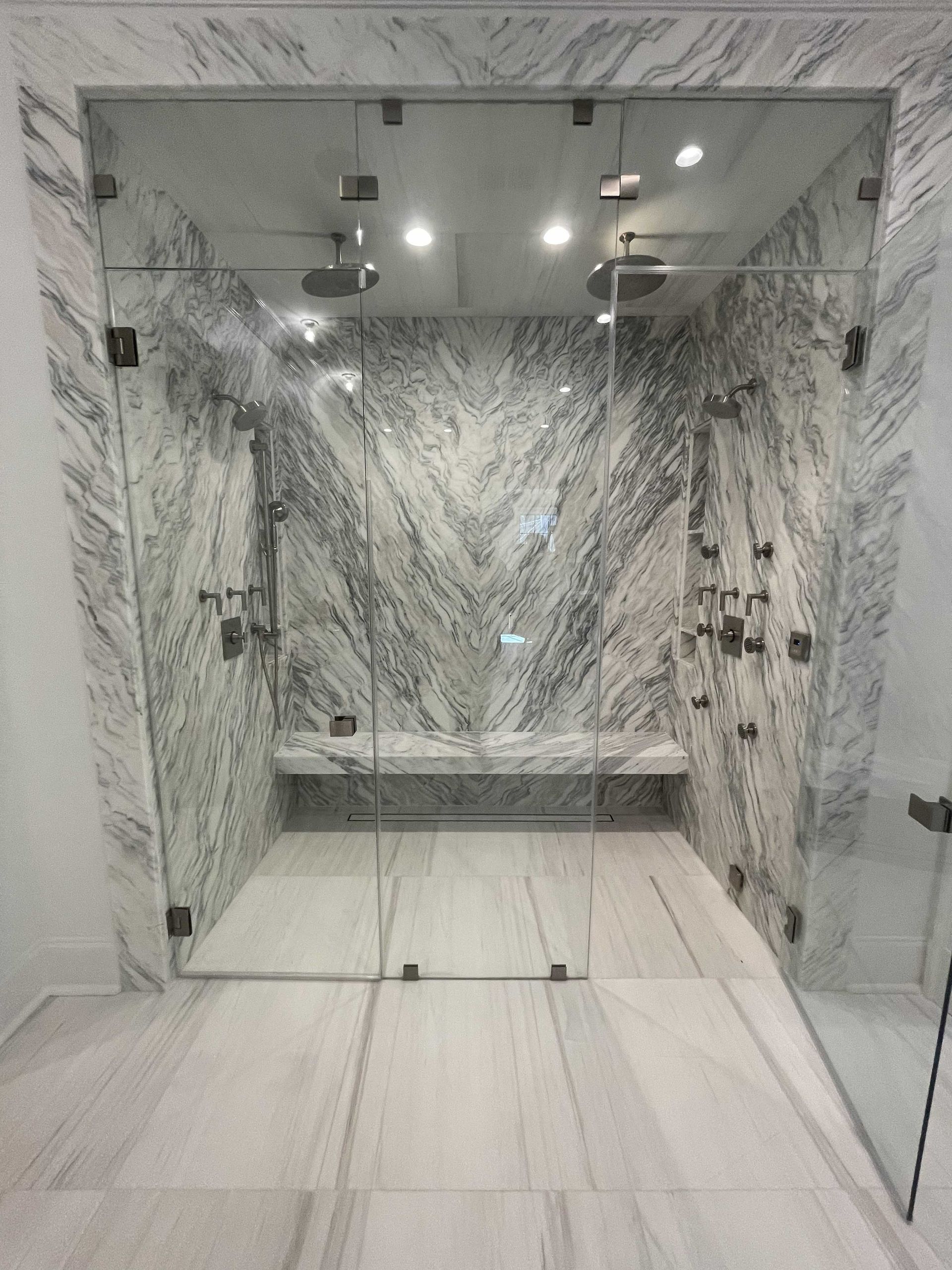 Luxurious marble shower with glass doors, multiple shower heads, and a bench.