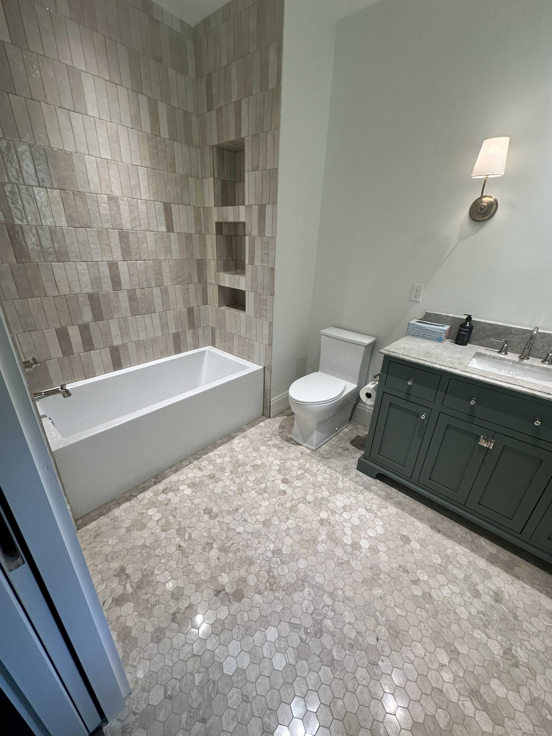 Bathroom with hexagon tile floor, white tub, gray tiled shower wall, green vanity, white toilet.