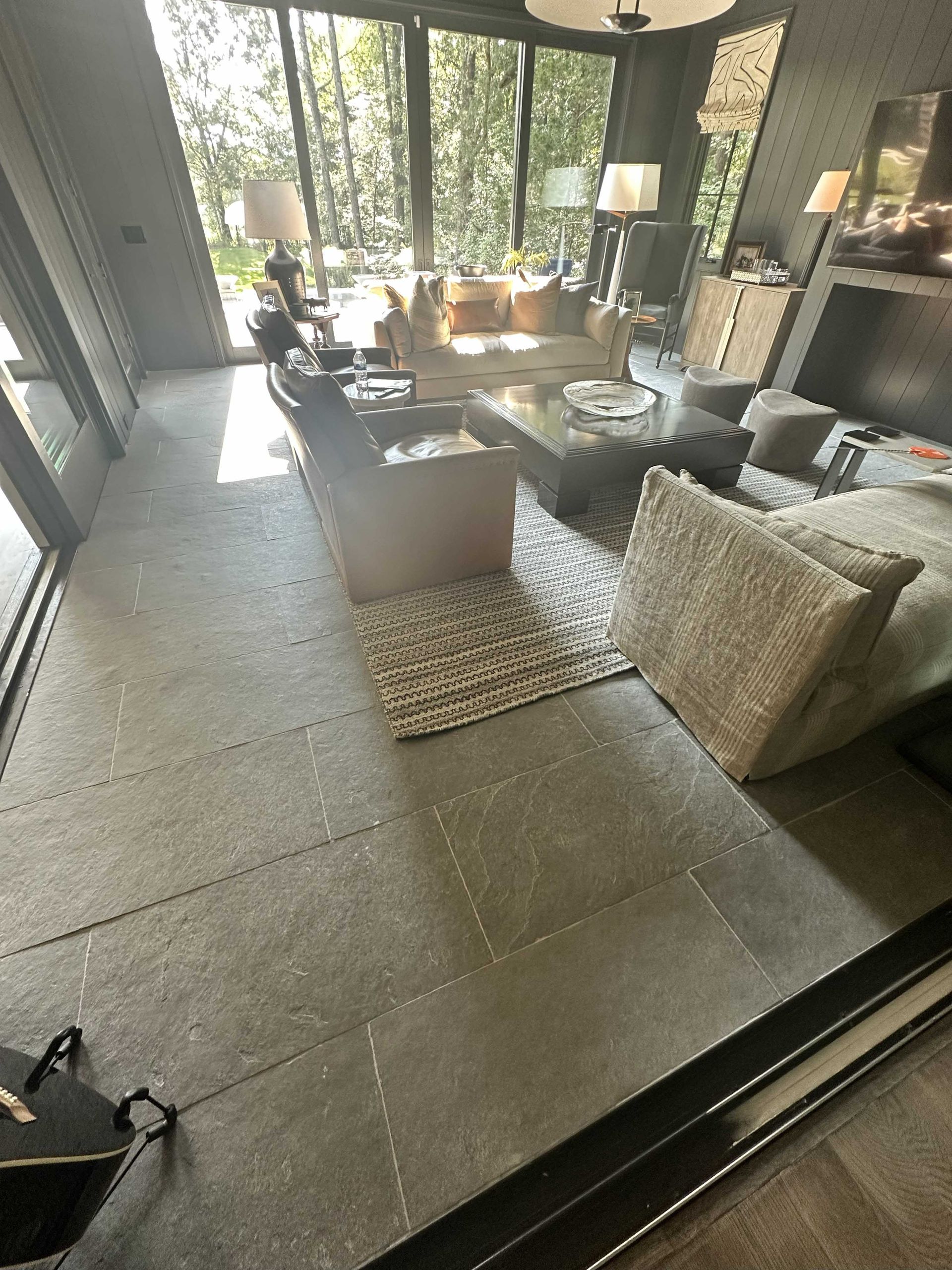 Living room with gray tiled floor, light-colored sofa, area rug, and windows overlooking trees.