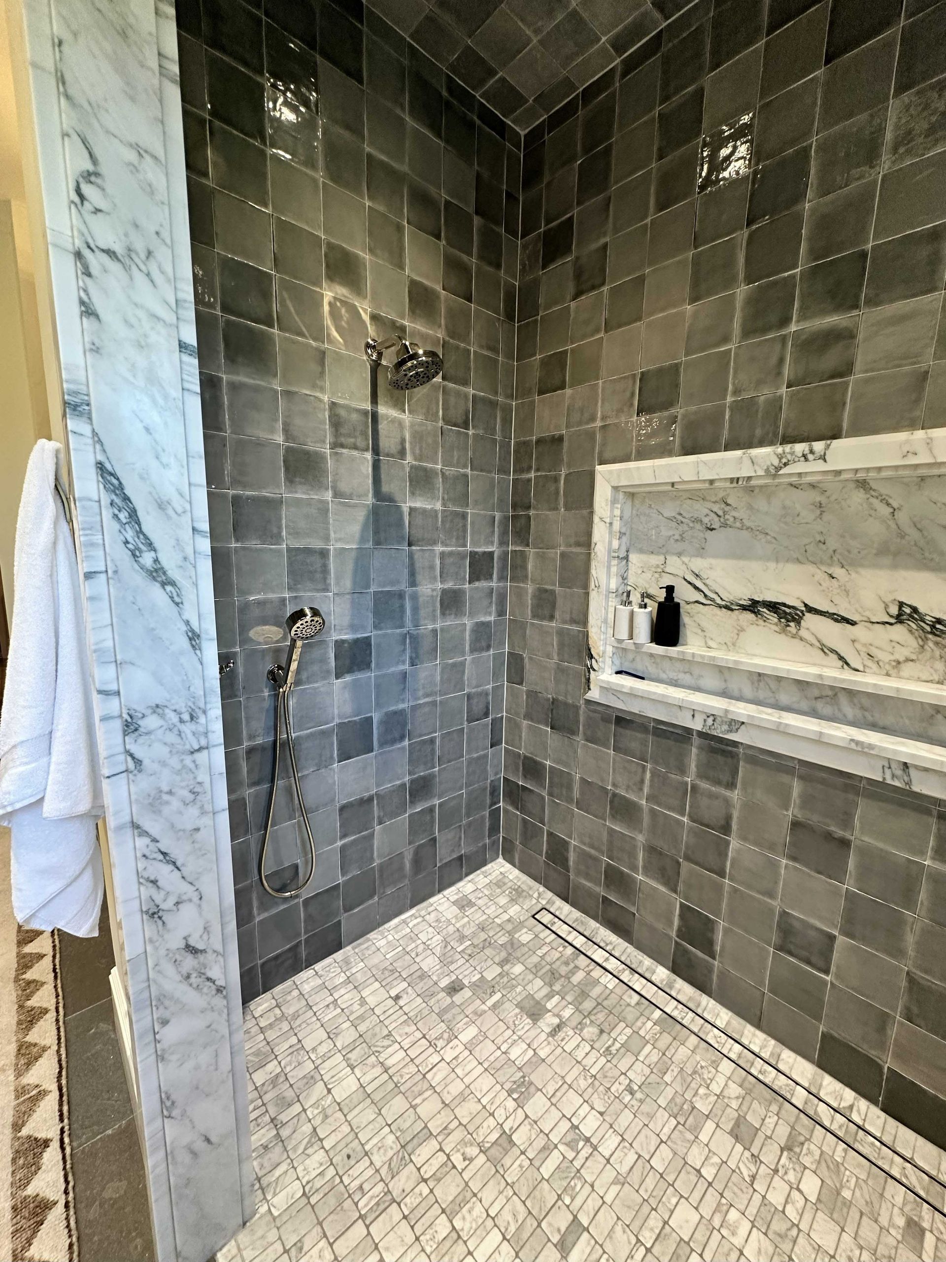 Shower with dark gray tile walls and a marble shelf and trim; small, white floor tiles.