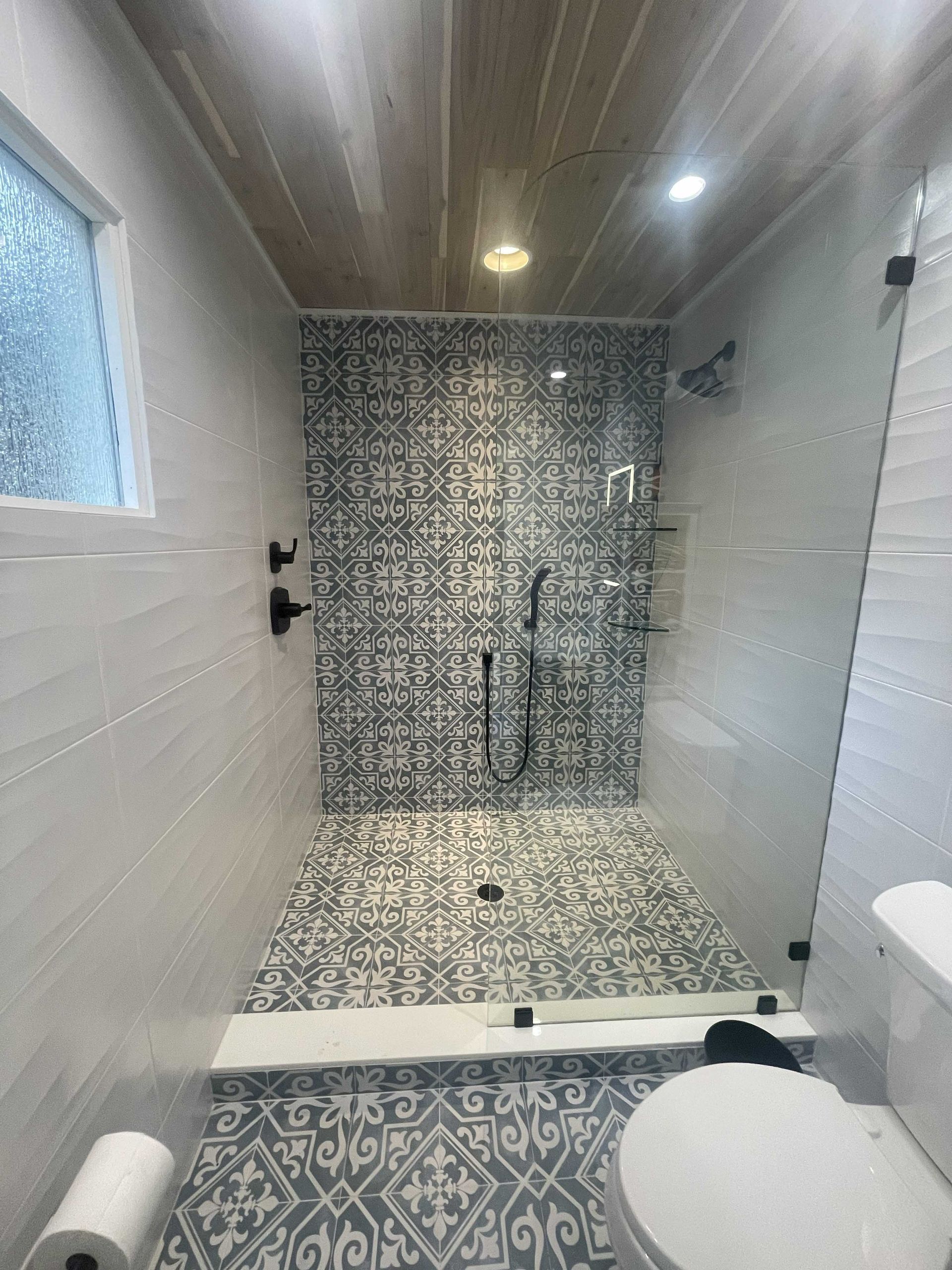 Bathroom with patterned tile shower, glass door, white walls, and wood ceiling.