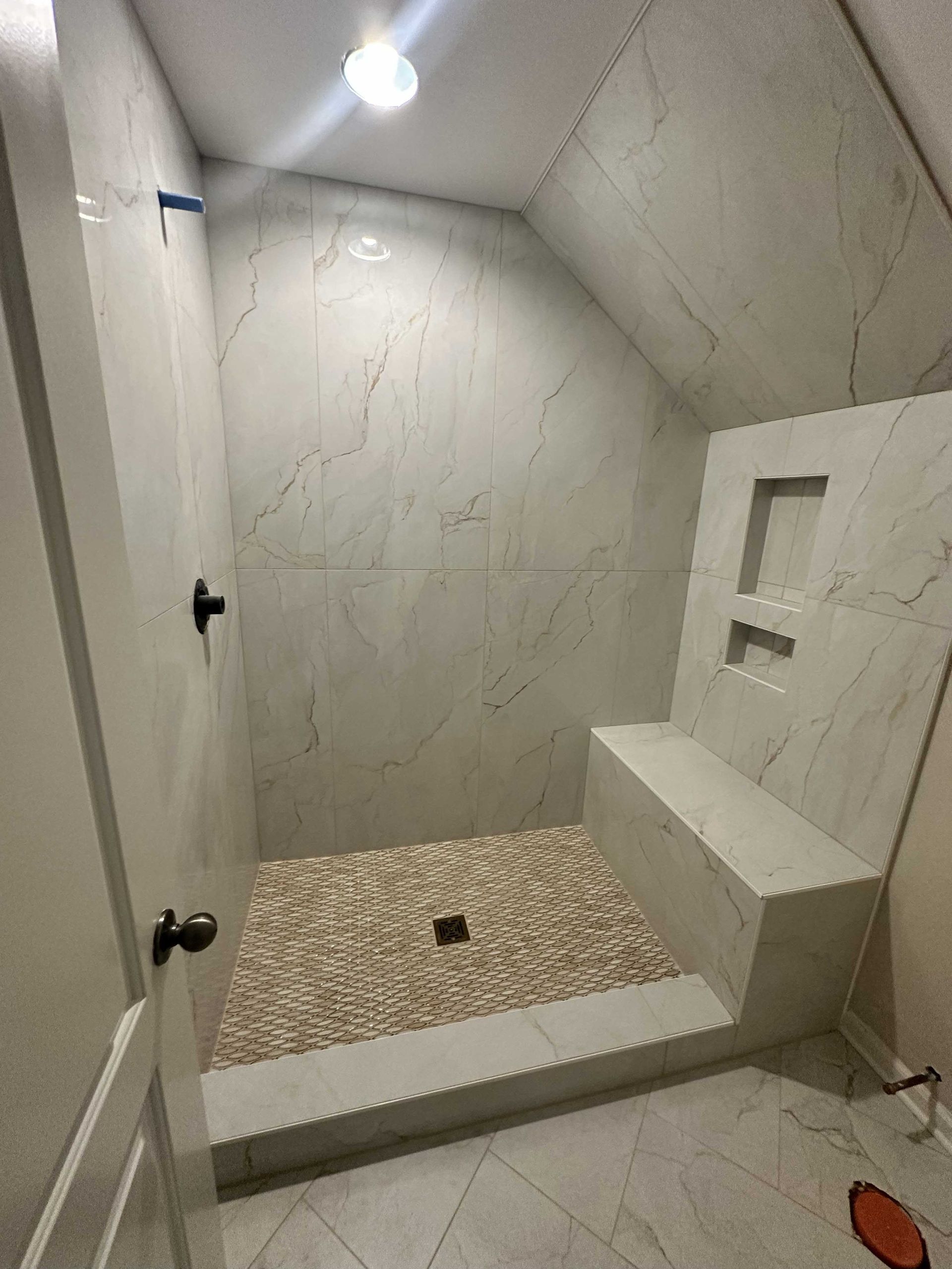 A newly tiled bathroom shower. White marble-like tiles and pebble-stone floor. Includes a built-in bench.