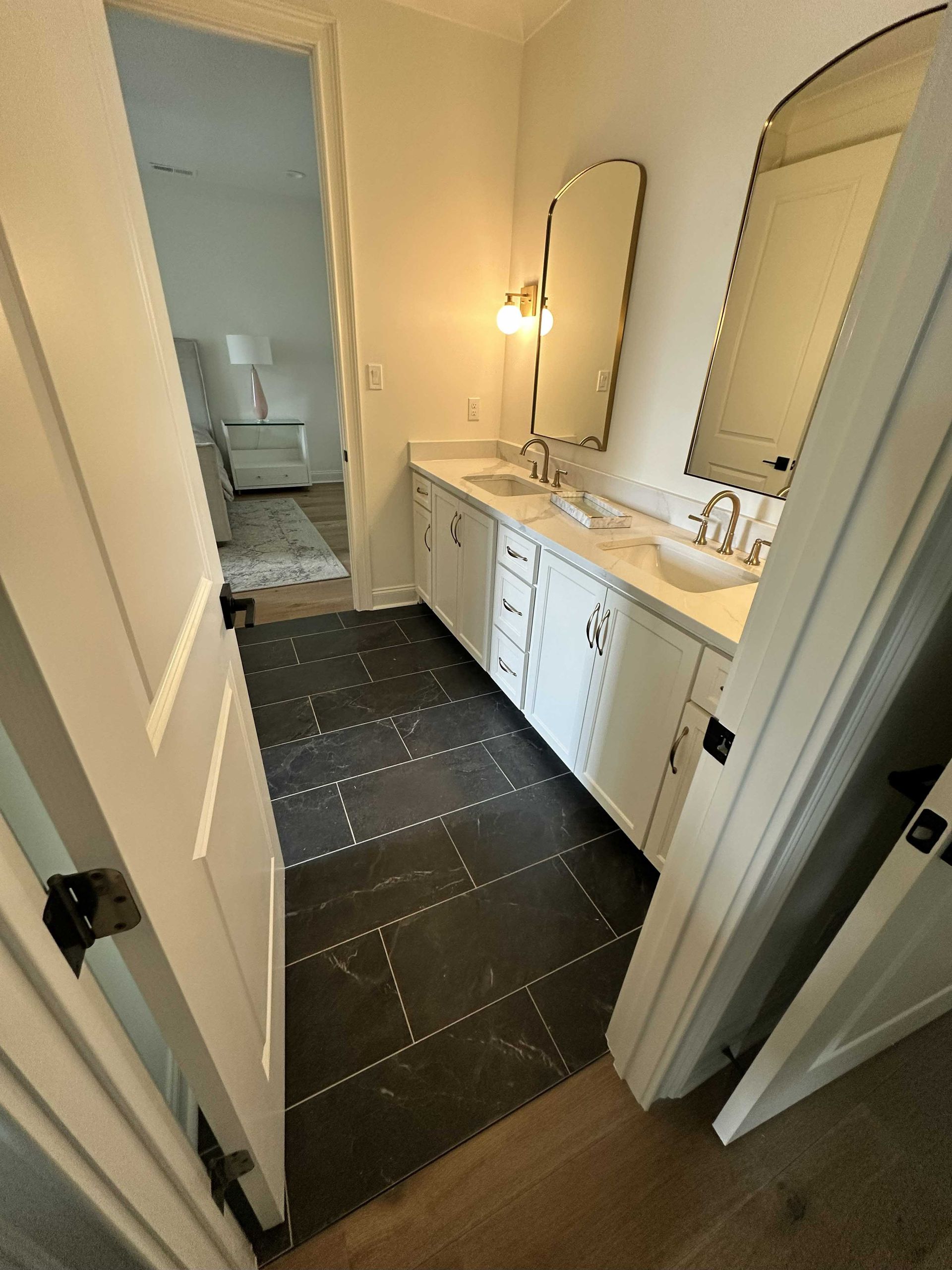 Bathroom with black tile floor, white cabinets, arched mirrors, and an open doorway to another room.