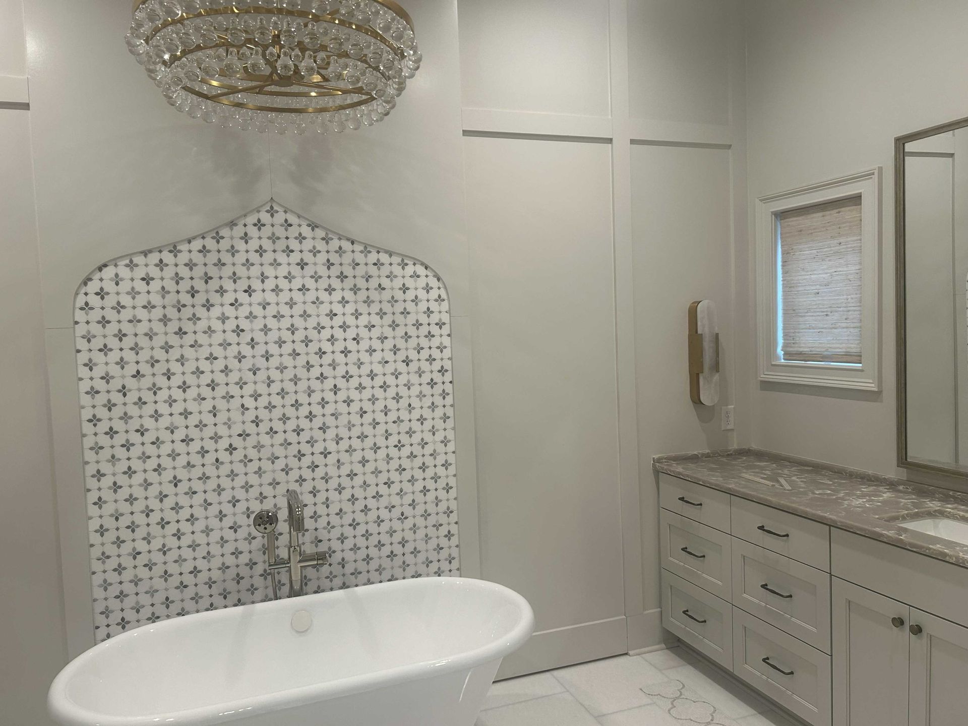 White bathroom with ornate tile wall behind a freestanding tub, chandelier, and vanity.