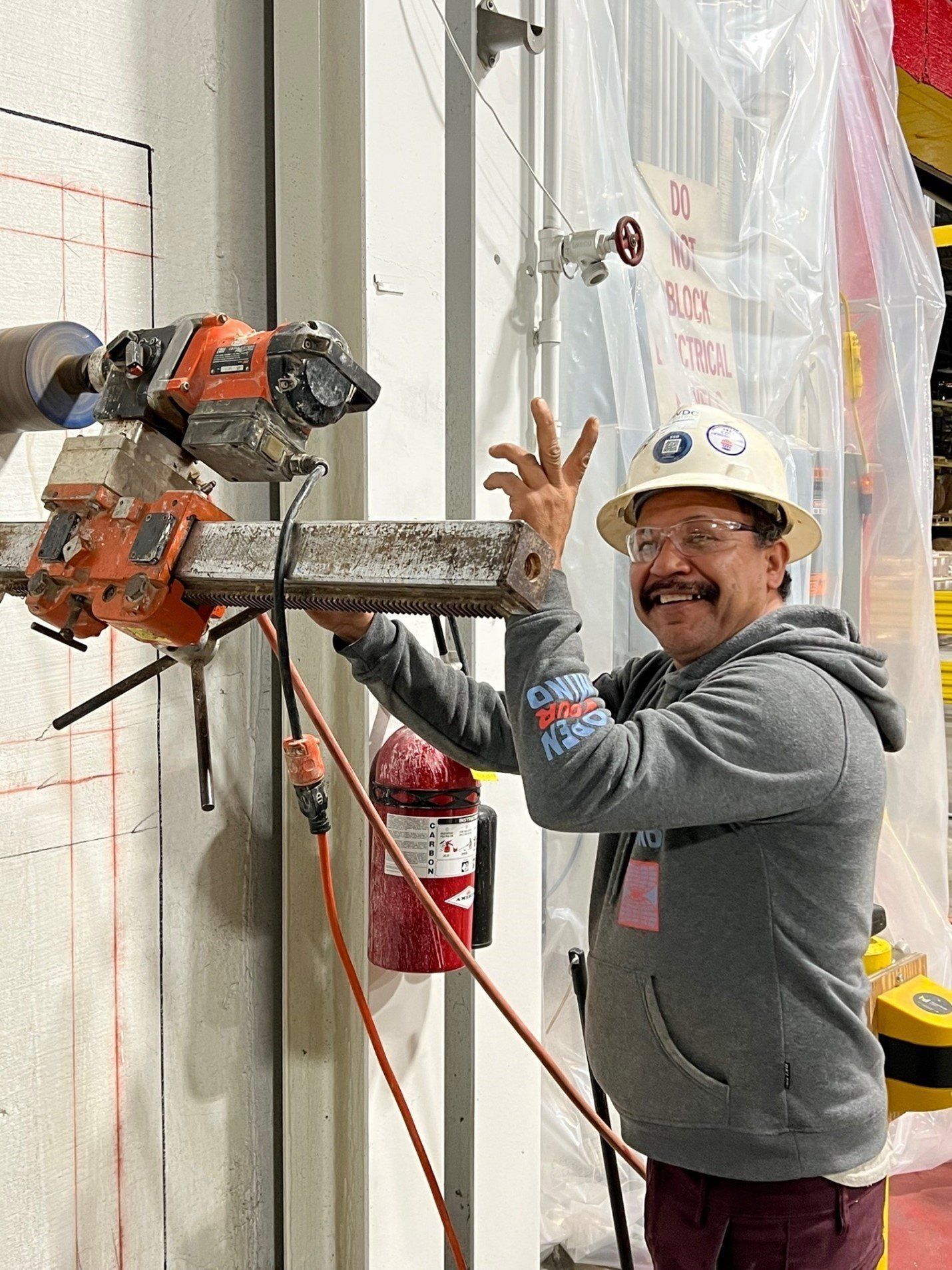Man in hard hat and safety glasses, smiling while working with a core drilling machine on a concrete wall.