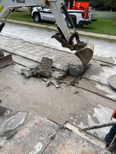 Bobcat excavator removing concrete from a street; other vehicles, cones and a person are present.