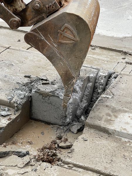 An excavator bucket breaks up concrete pavement, revealing a trench.
