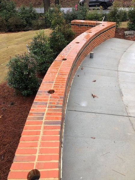 Curving brick wall with evenly spaced holes, alongside a concrete path and greenery.
