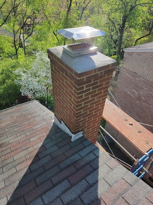 Brick chimney with metal cap on a shingled roof, with trees and bright sky in the background.
