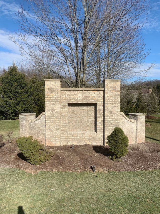 Brick entrance sign with brown and tan bricks, with two small bushes, and a leafless tree behind.