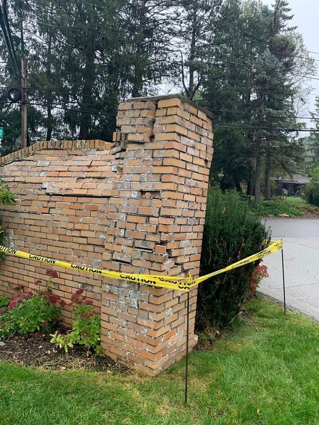 Damaged brick wall with yellow caution tape in front of a suburban yard, possibly due to weather.