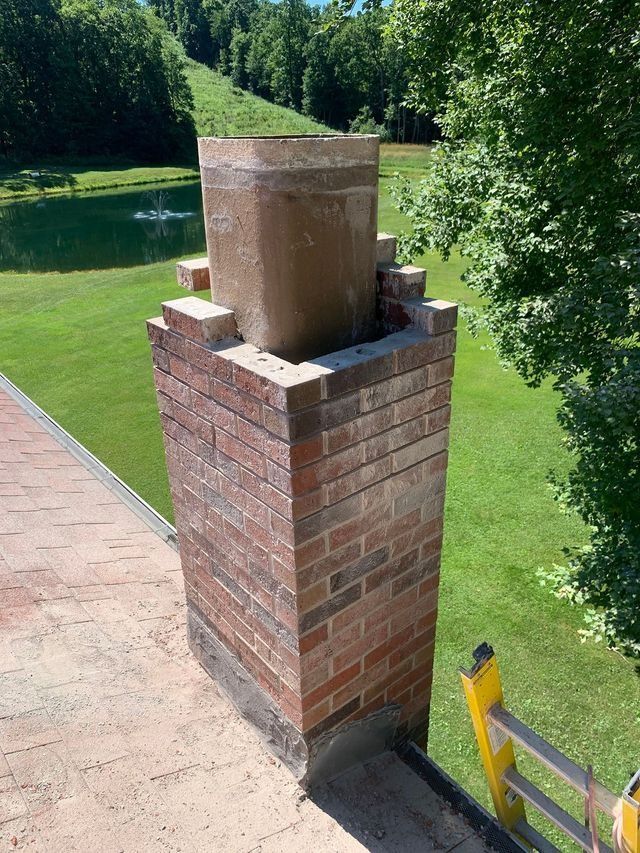Brick chimney on a roof with a concrete top and a green grassy background.