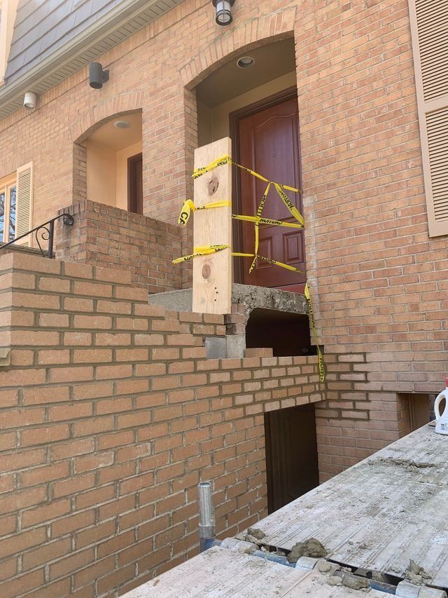 Damaged brick entrance to a building, with yellow tape blocking access. A wooden plank supports the door.