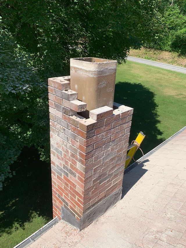 Brick chimney on a rooftop; new brickwork and concrete flue visible, surrounded by trees and grass.