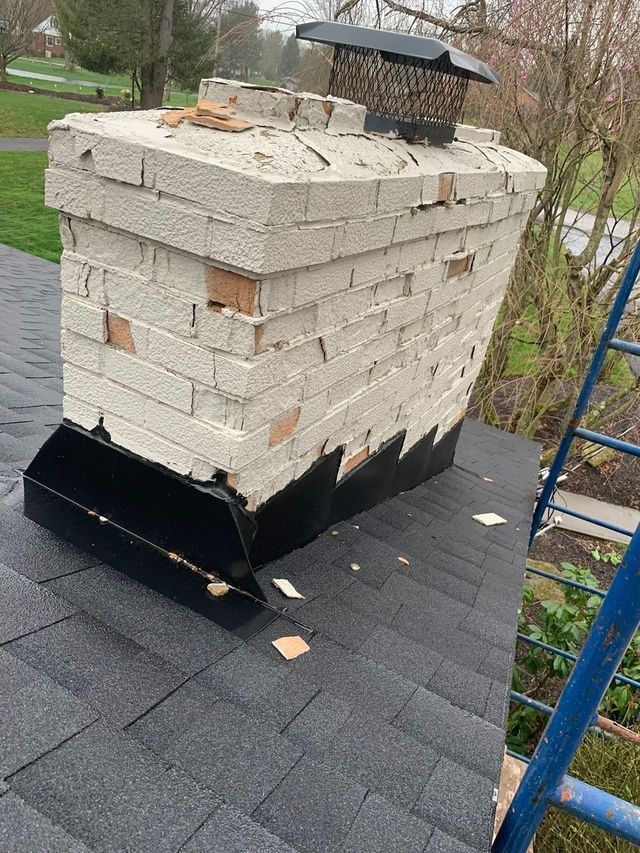 Damaged chimney on a dark shingled roof. White bricks are crumbling. Black metal flashing is present.