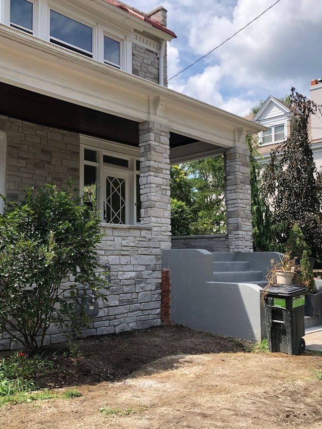 Stone facade of a house with a porch and gray steps. Lush greenery and cloudy sky.
