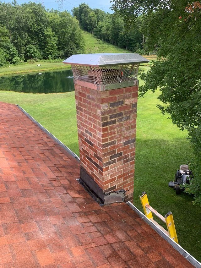 Brick chimney on a red shingled roof, with a metal cap. A ladder and lawn are visible in the background.