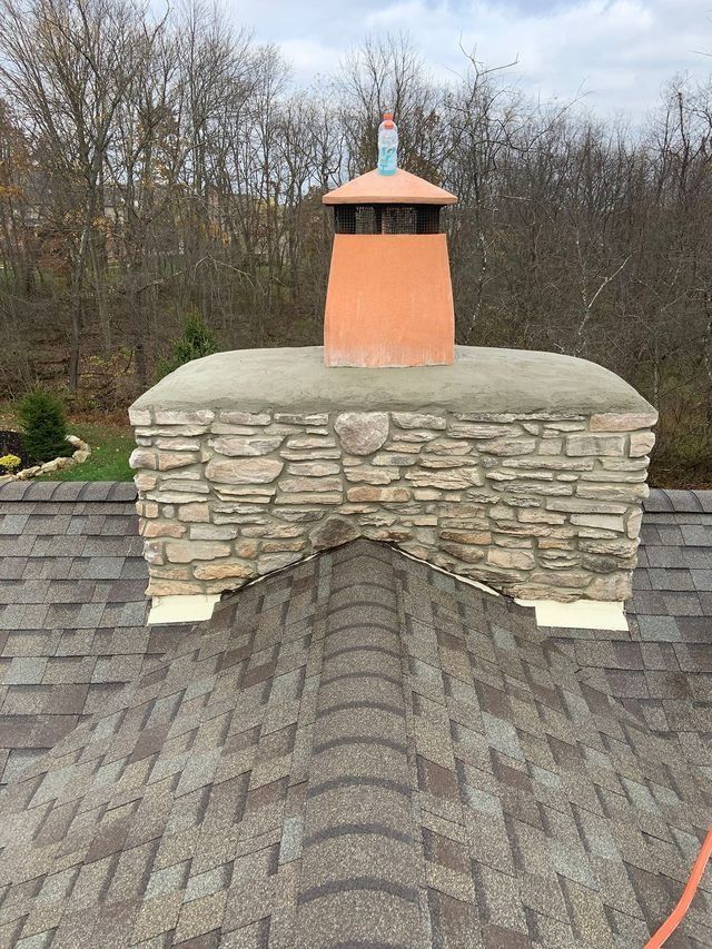 Stone chimney on a brown shingled roof, with an orange cap, set against a cloudy sky.