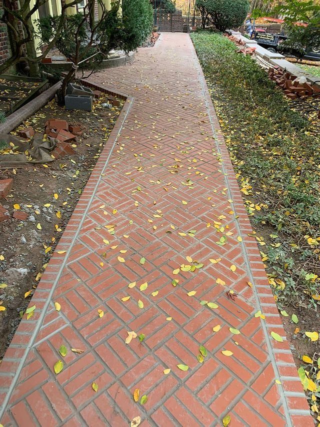 Brick pathway lined with grass and trees; fallen yellow leaves scattered across the bricks.