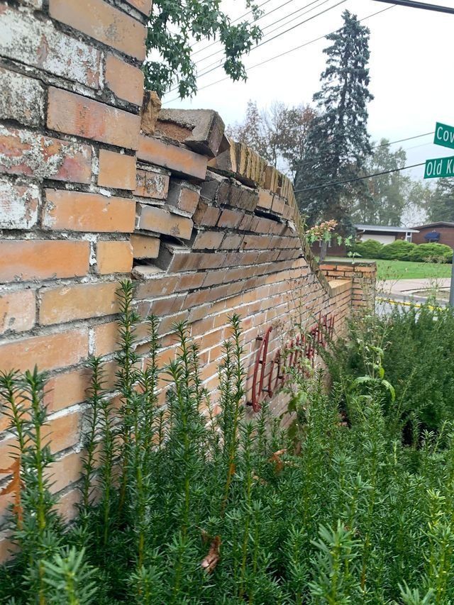 Brick wall with significant damage, leaning and crumbling, overgrown with green plants. Street corner in background.
