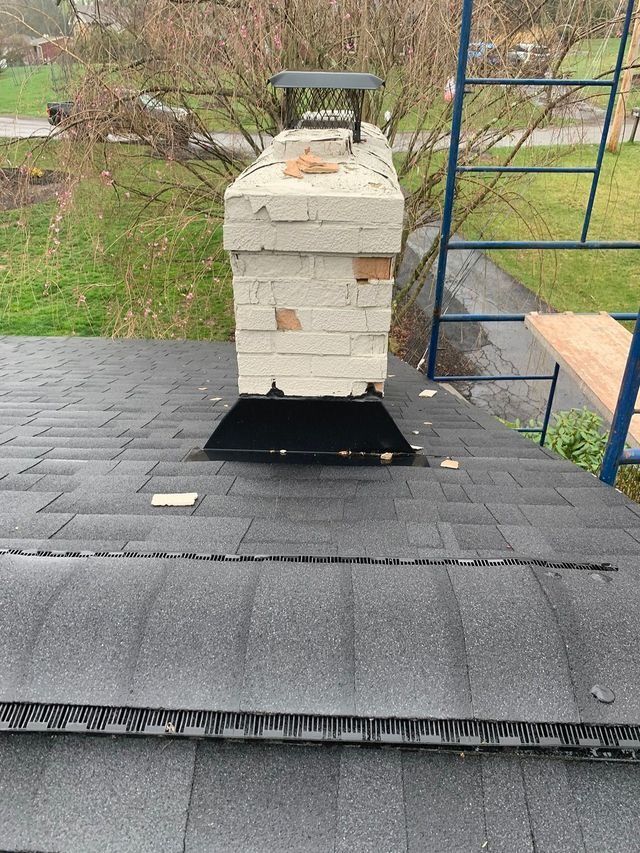 Chimney on a dark shingled roof. Brick chimney shows damage. Scaffolding and overcast sky in the background.