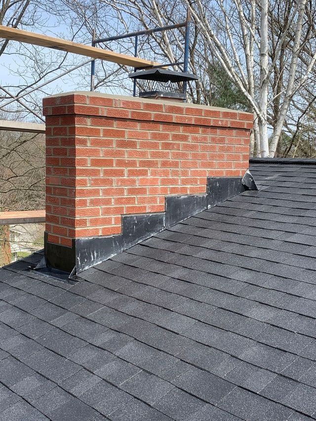 Brick chimney on a dark-shingled roof. Black flashing provides a seal. Scaffolding in the background.