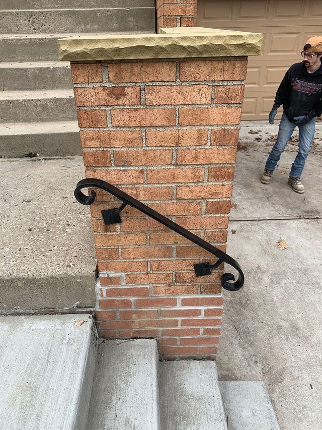 Brick pillar with black handrail next to concrete steps, a person in motion in the background.