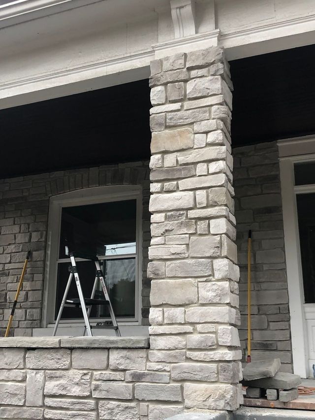 Stone-clad porch column with a gray-stone veneer, supporting a white trim. A ladder sits in front of a window.