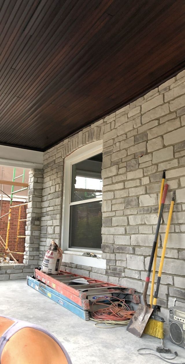Porch with stone wall, dark ceiling, tools leaning against it; construction in background.