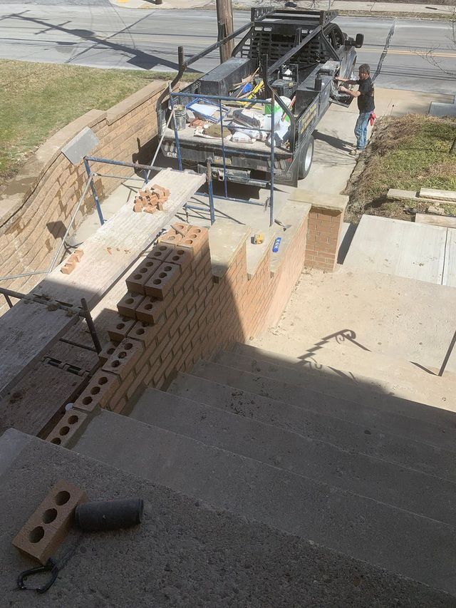 Bricklayers building a low wall next to a concrete staircase. A truck with equipment parked nearby.