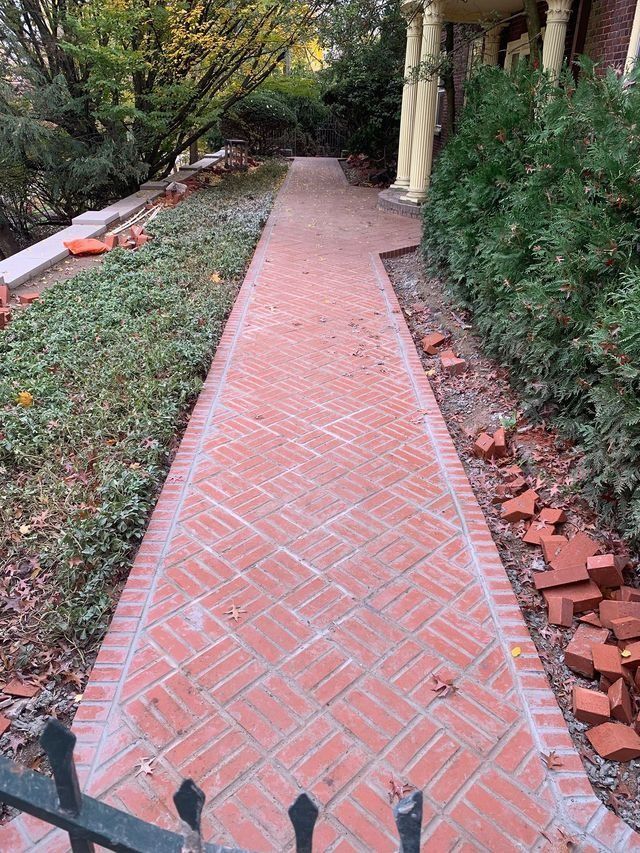 Brick walkway with brick border next to green landscaping and building. Bricks in a herringbone pattern.