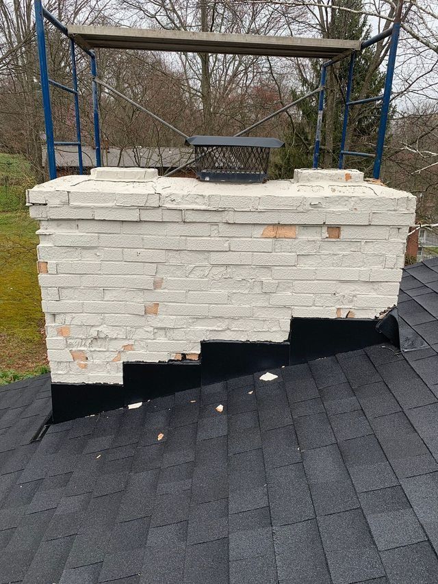 Damaged brick chimney with missing sections, resting on a black asphalt shingle roof. Scaffolding in background.