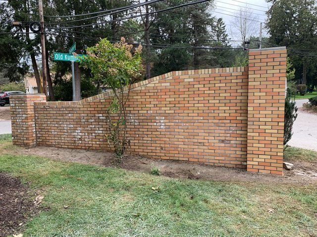 Brick wall with columns in front of a green street sign; a small tree grows in front.