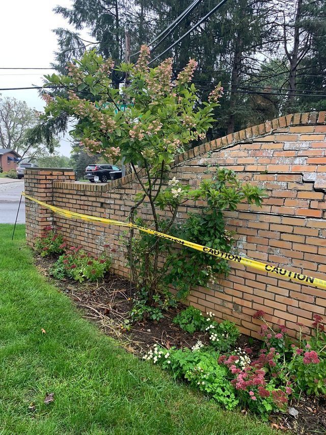 Brick wall with yellow caution tape, leaning, and plants growing in front.