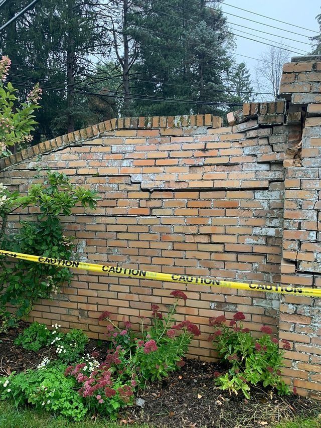 Brick wall with significant cracks, caution tape across, and a flower bed in front.