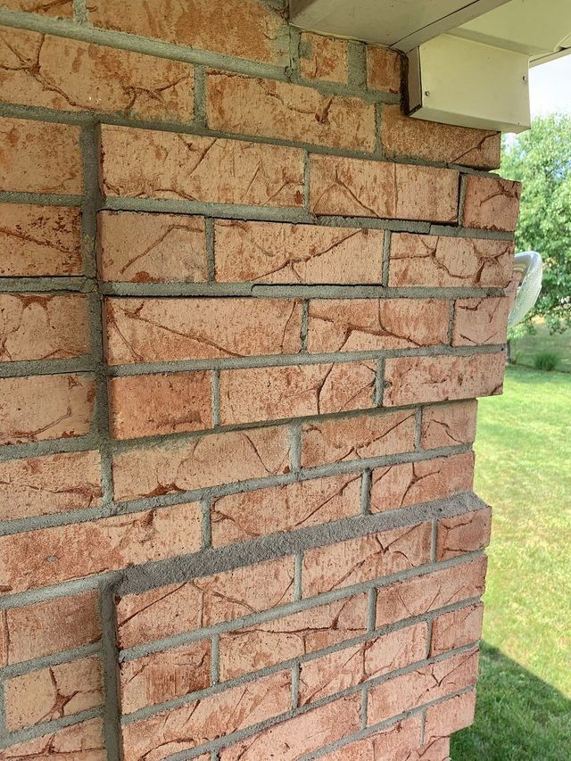 Red brick wall with displaced bricks, potentially due to structural issues, set outdoors with greenery visible.