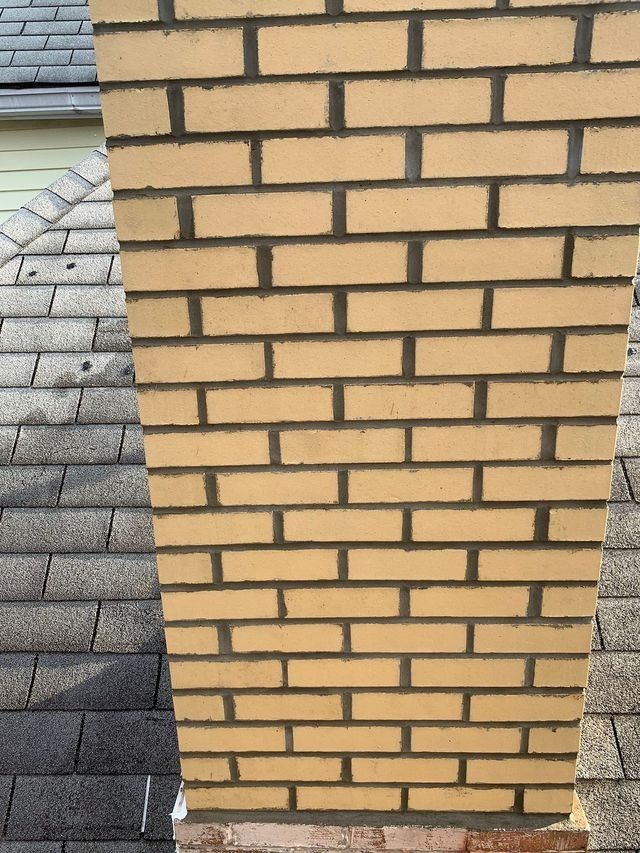 Brick chimney with dark mortar, atop a gray shingle roof, against a sunny sky.