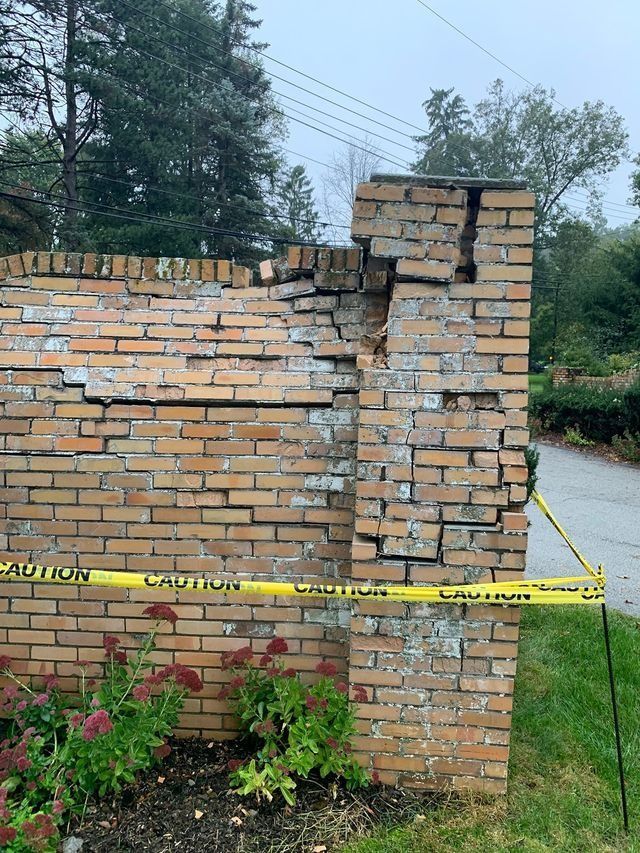 Damaged brick wall section, cracked bricks, caution tape, green grass, plants, overcast sky.