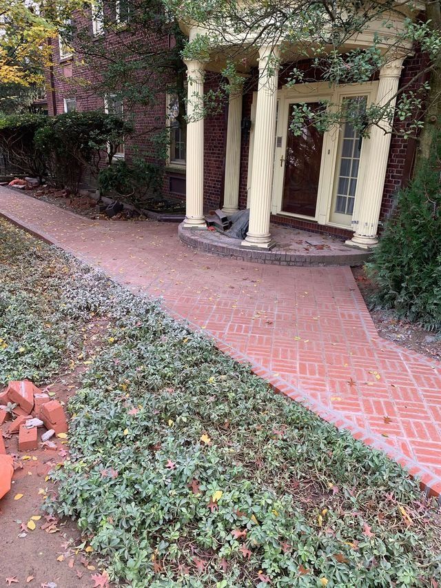 Brick pathway leading to a house with columns; foliage and fallen bricks are along the path.