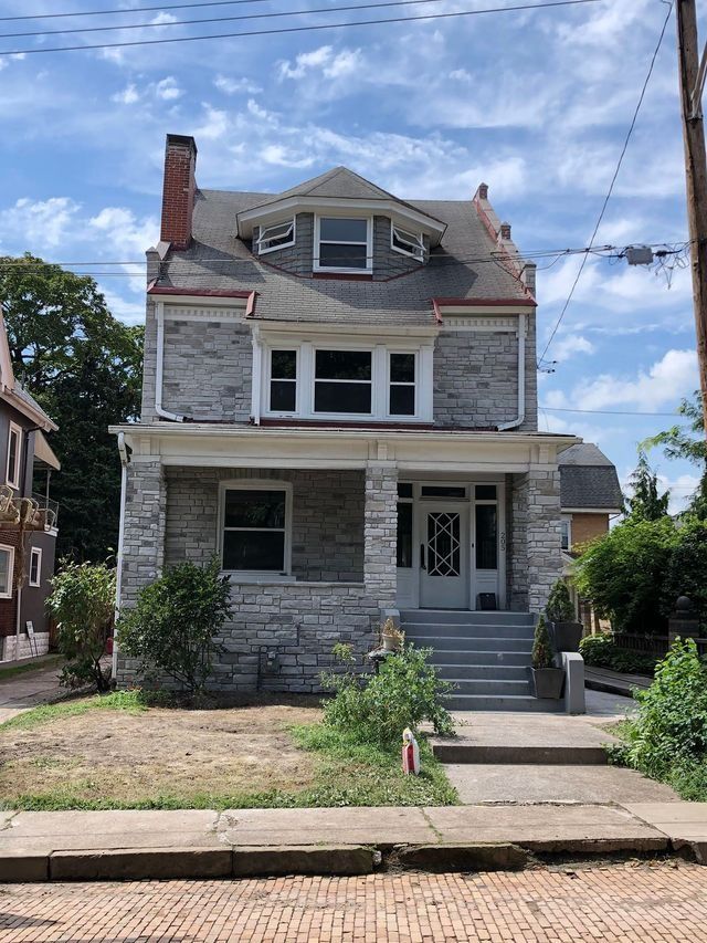 Two-story gray stone house with porch and steps, a front door with decorative glass, and a red-brick chimney.