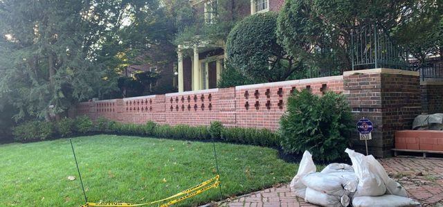 A brick wall with decorative cutouts in front of a house. Green grass and foliage are in view.