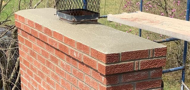 Brick chimney with a concrete top and metal chimney cap, with a wooden board nearby.
