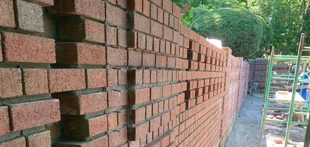 Brick wall construction, showing laid bricks with a ladder and greenery in the background.