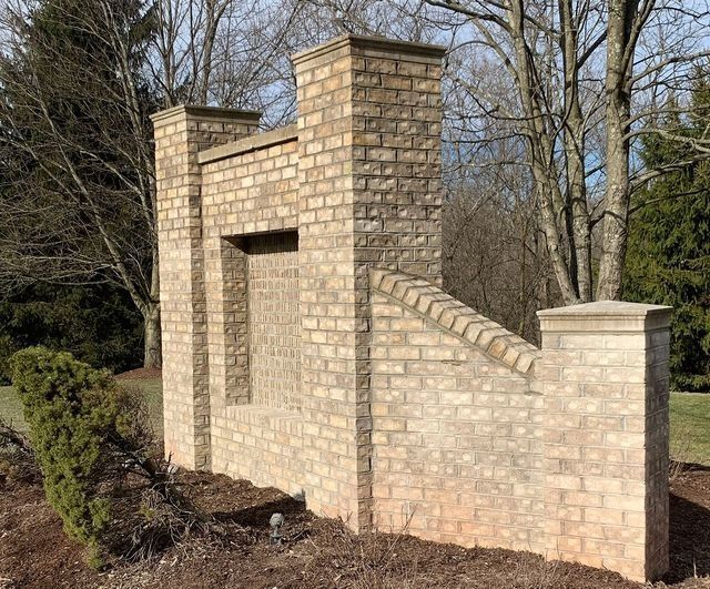 Brick entrance wall with columns and a decorative window-like feature. Set in front of trees and grass.