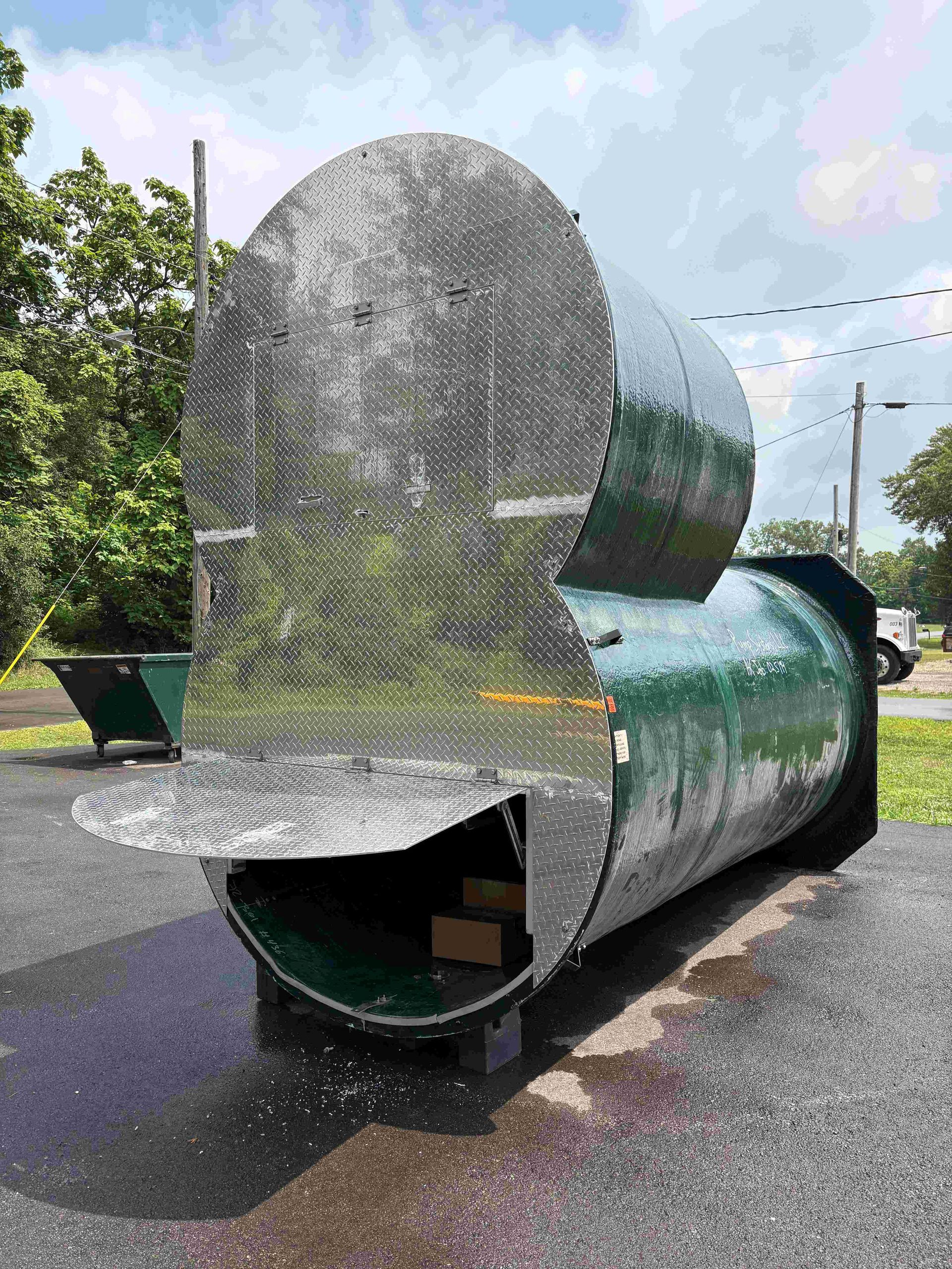Large silver metal sculpture outdoors, with a curved reflective surface and green base on pavement.