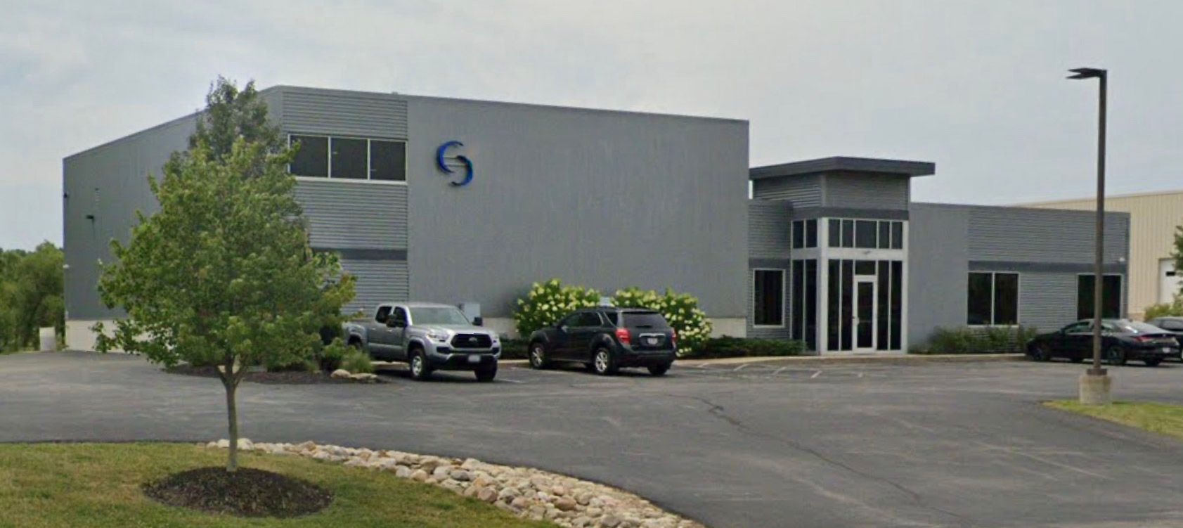 Gray industrial building with cars parked out front and a tree in the foreground.