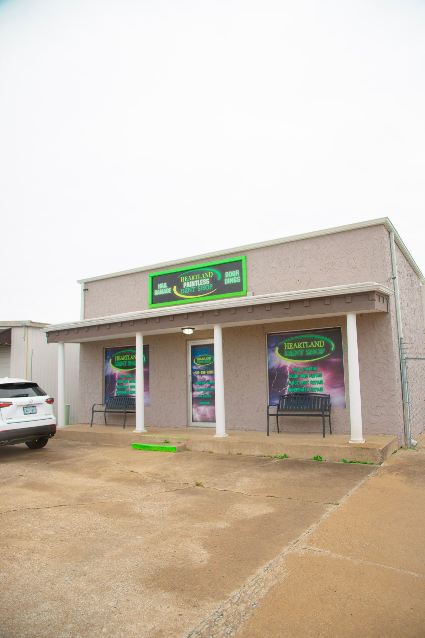 A small brick building with green signage and large posters, two benches in front; a white SUV parked to the left.