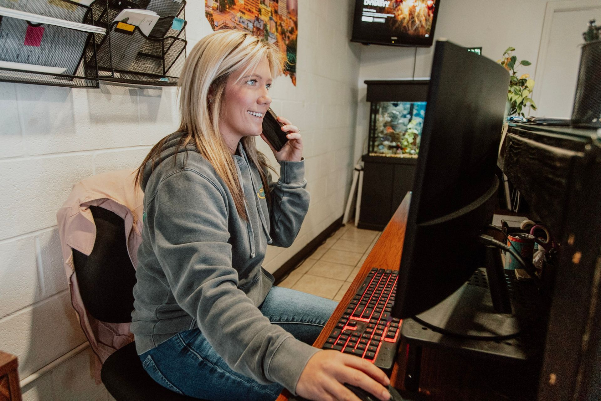 Woman in gray hoodie on the phone at a desk, typing on a red-lit keyboard. A computer monitor and fish tank are visible.
